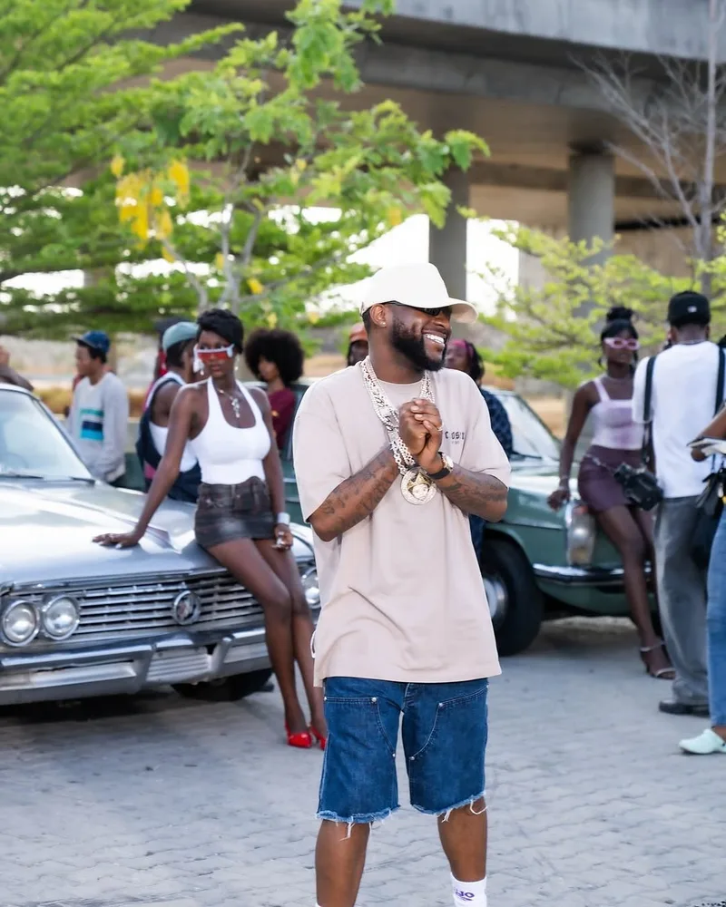 Cassper Nyovest souriant en casquette blanche sur un tournage, voitures vintage et figurants derrière lui