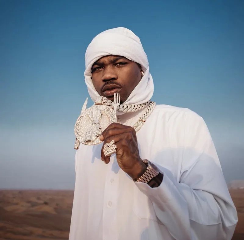 Gunna en tenue blanche dans le désert, tenant un gros pendentif diamant, montre au poignet