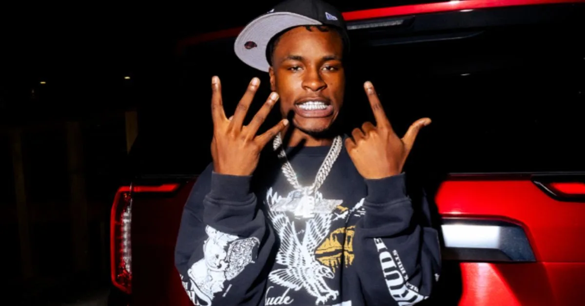 Le rappeur Mudbaby Ru, casquette et chaînes, faisant des signes de la main devant une voiture rouge